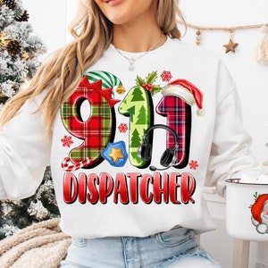 Puede incluir: Sudadera blanca con un diseño festivo de Despachador del 911. El diseño presenta los números 911 en patrones de cuadros, un gorro de Papá Noel y unos auriculares. La palabra "DISPATCHER" está en rojo. También se ve una taza con Papá Noel.