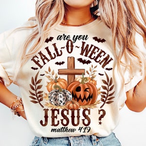May include: A white t-shirt with a graphic design featuring a cross with pumpkins and bats. The text reads "Are you Fall-O-Ween Jesus? Matthew 4:19".