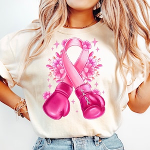 May include: A white t-shirt with a pink ribbon and boxing gloves design. The ribbon is surrounded by pink flowers and sparkles.