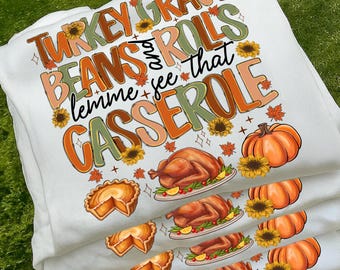 May include: White t-shirts stacked, each featuring a Thanksgiving-themed graphic. The design includes text that reads "Turkey Gravy Beans and Rolls lemme see that Casserole" with images of a turkey, pie, and pumpkins.