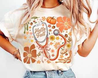 May include: A white t-shirt with a stethoscope, pumpkins, and flowers in orange, brown, and pink colors. The text "Hello Fall" is written on the stethoscope.