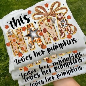 May include: A stack of white t-shirts with the text "this NANA loves her pumpkins." The word "NANA" is decorated with fall-themed patterns, including leaves, acorns, and a star. The design is in shades of orange, brown, and white.
