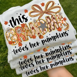 May include: A stack of white sweatshirts with the text "this GRANDMA loves her pumpkins" in a fall-themed design. The design includes orange leaves, acorns, and a brown bow.