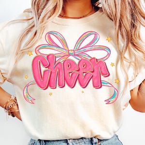 May include: Cream-colored sweatshirt with a large pink "Cheer" graphic. The word "Cheer" is in a bubble letter style, outlined in a darker pink. A colorful bow and ribbon design surrounds the word, with small yellow stars scattered around.