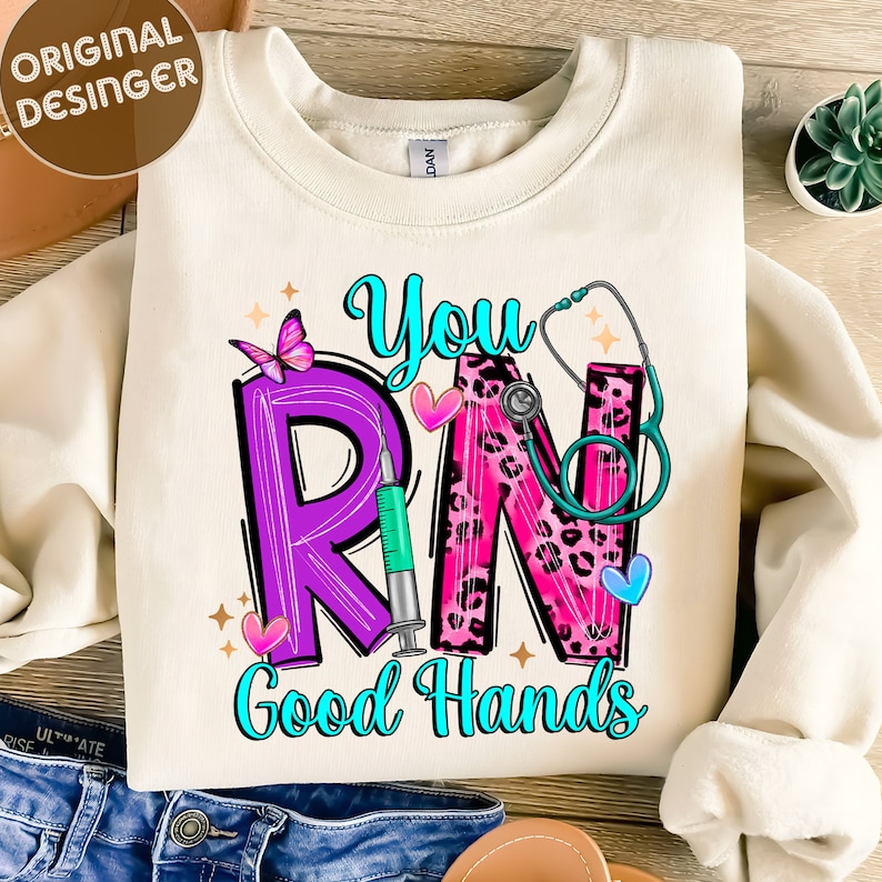 May include: A white sweatshirt with a pink, purple, and teal graphic design that says "You RN Good Hands". The graphic includes a stethoscope, a butterfly, a syringe, and hearts.