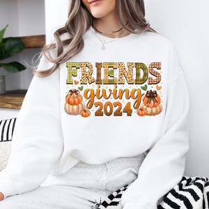 May include: White sweatshirt with the text "FRIENDS giving 2024" in brown and green letters with a plaid and leopard print design. The text is surrounded by small hearts and two illustrations of pumpkins.