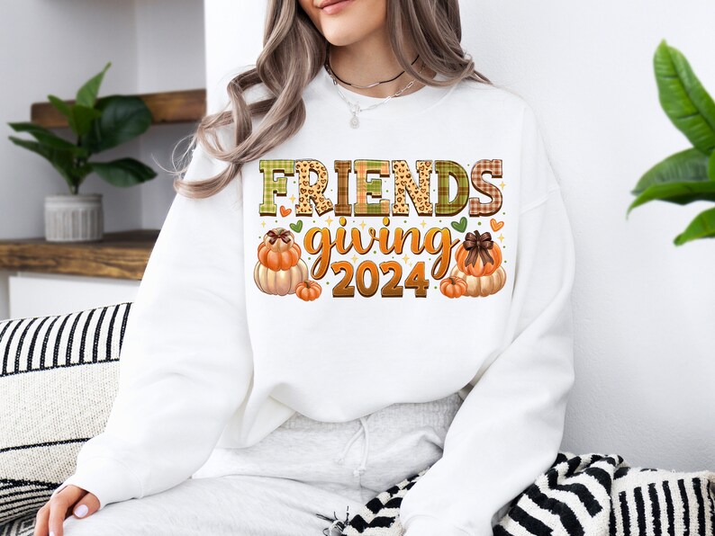 May include: White sweatshirt with the text "FRIENDS giving 2024" in brown and green letters with a plaid and leopard print design. The text is surrounded by small hearts and two illustrations of pumpkins.