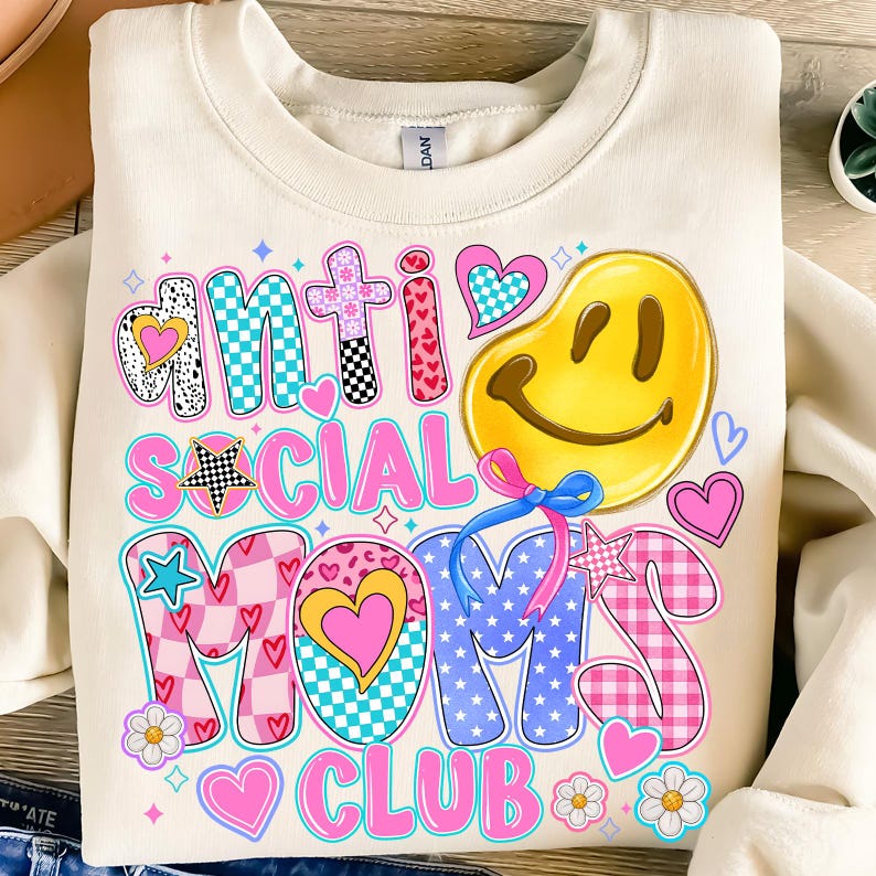 May include: A white sweatshirt with a colorful graphic design that says "Anti Social Moms Club". The design features a smiley face, hearts, stars, and checkered patterns.