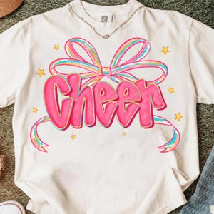 May include: White t-shirt with a pink "Cheer" graphic in a bubble letter font, accented by a colorful bow and stars. The shirt has a crew neck and short sleeves. The text and bow are outlined in a darker pink.