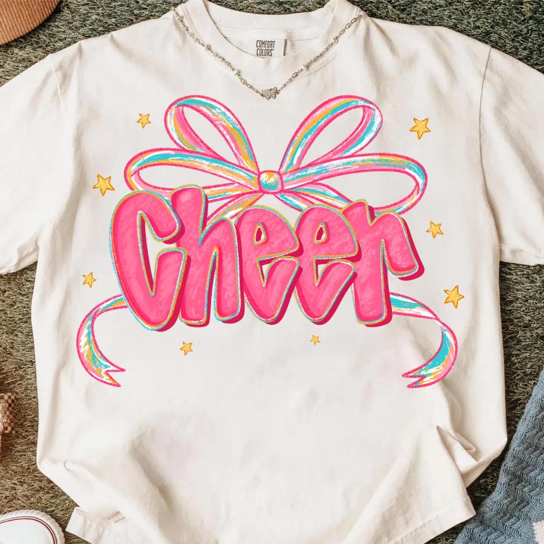 May include: White t-shirt with a pink "Cheer" graphic in a bubble letter font, accented by a colorful bow and stars. The shirt has a crew neck and short sleeves. The text and bow are outlined in a darker pink.