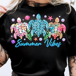 May include: Black t-shirt with a graphic of three colorful sea turtles, seashells, and starfish. The text "Summer Vibes" is printed below the turtles.