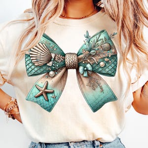 May include: A white t-shirt with a large, teal and brown bow with seashells, starfish, and floral accents. The bow is tied with a rope and has a mermaid-like scale pattern.