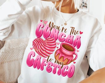 May include: White sweatshirt with pink lettering that says "You're the Concha to my Cafecito" with a pink heart and a cartoon image of a concha pastry and a cup of coffee.