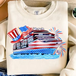 May include: Cream-colored sweatshirt featuring a patriotic cruise ship design. The ship is decorated with red, white, and blue stripes and stars, with American flags and a top hat. Fireworks are depicted in the background.