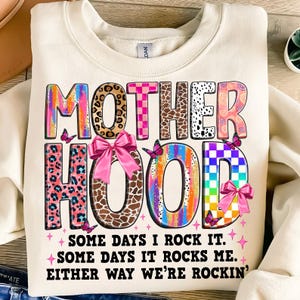 May include: A white sweatshirt with a colorful graphic that says "MOTHERHOOD" with a pink bow and butterflies. The text below the graphic says "Some days I rock it. Some days it rocks me. Either way we're rockin'."