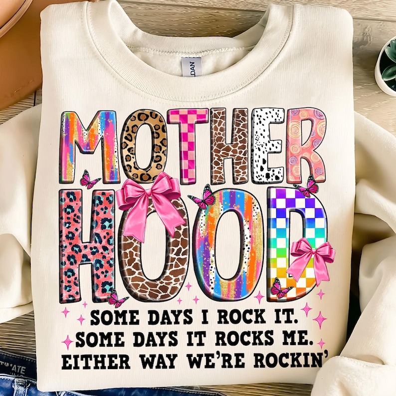 May include: A white sweatshirt with a colorful graphic that says "MOTHERHOOD" with a pink bow and butterflies. The text below the graphic says "Some days I rock it. Some days it rocks me. Either way we're rockin'."