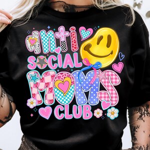 May include: Black t-shirt with a colorful graphic design that says "Anti Social Moms Club". The graphic features a smiley face, hearts, stars, and checkered patterns.