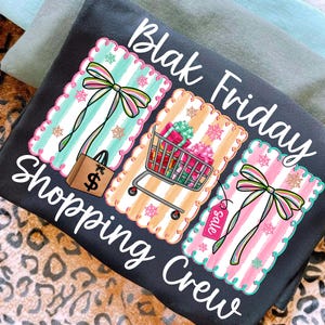 May include: Dark gray t-shirt with the words "Blak Friday Shopping Crew" in white script. The shirt features three decorative panels with shopping-themed graphics, including a shopping bag, shopping cart, and sale tag.