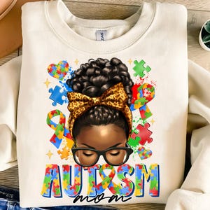 May include: White sweatshirt with a colorful graphic of a Black woman with a bun and a leopard print bow. The graphic includes puzzle pieces, hearts, and the words "Autism Mom".
