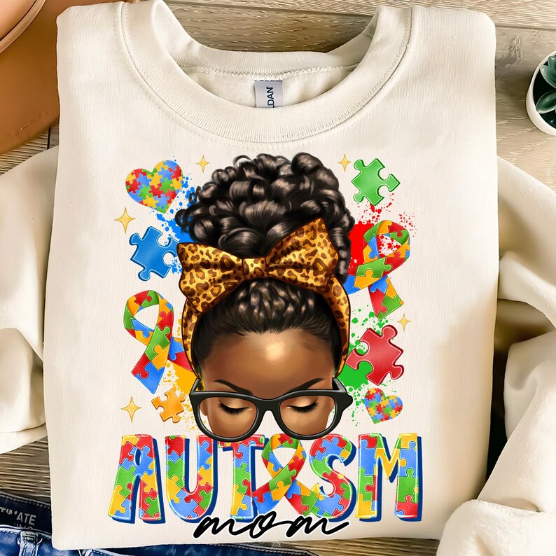 May include: White sweatshirt with a colorful graphic of a Black woman with a bun and a leopard print bow. The graphic includes puzzle pieces, hearts, and the words "Autism Mom".