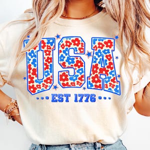 May include: A white t-shirt with a blue and red floral pattern spelling out "USA" with the text "...EST 1776..." underneath.