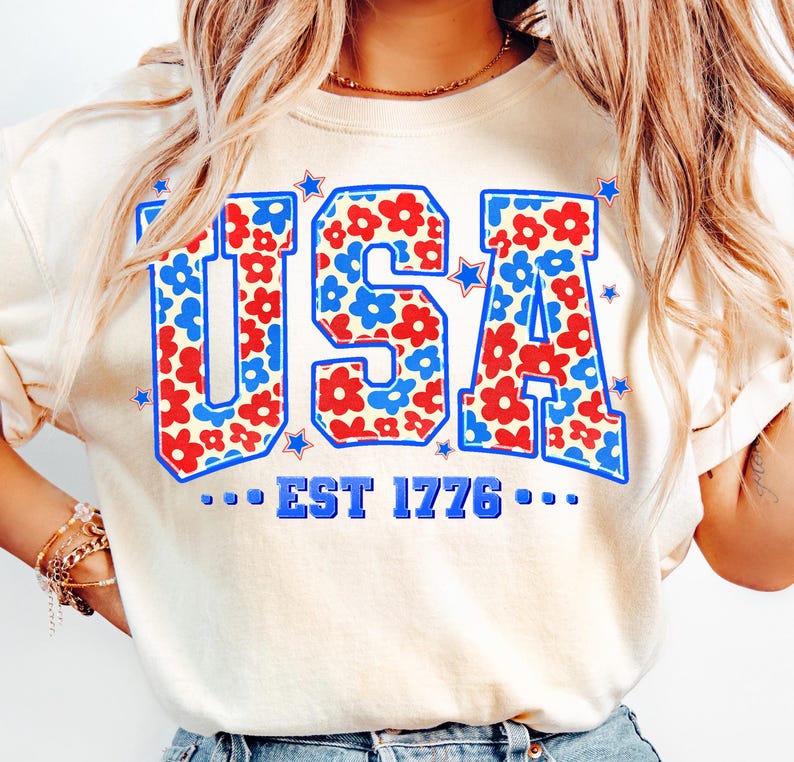May include: A white t-shirt with a blue and red floral pattern spelling out "USA" with the text "...EST 1776..." underneath.