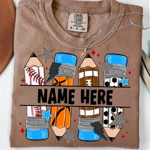 Pode incluir: Camiseta marrom com um design de l&aacute;pis com gr&aacute;ficos de beisebol, basquete, futebol americano e bola de futebol. O texto "NAME HERE" &eacute; impresso no centro. A camisa tamb&eacute;m tem uma etiqueta Comfort Colors.