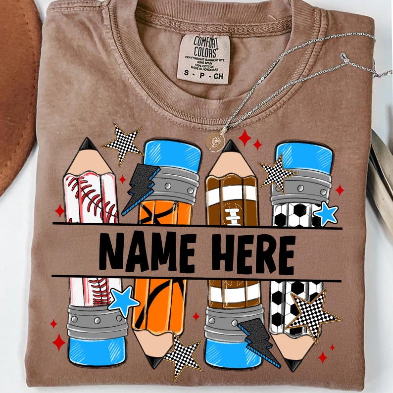Pode incluir: Camiseta marrom com um design de l&aacute;pis com gr&aacute;ficos de beisebol, basquete, futebol americano e bola de futebol. O texto "NAME HERE" &eacute; impresso no centro. A camisa tamb&eacute;m tem uma etiqueta Comfort Colors.