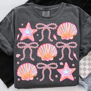 May include: A dark grey t-shirt with a colourful design featuring seashells, starfish, and pearl bows. The design is repeated throughout the shirt.