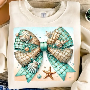 May include: Cream-colored sweatshirt featuring a watercolor design of a teal and brown bow adorned with seashells, pearls, and a starfish. The bow is set against a beach scene with a light blue sky.