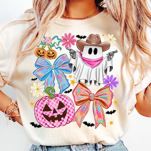 May include: A cream-coloured t-shirt with a Halloween design. The graphic features a ghost in a cowboy hat holding guns, alongside pumpkins, bows, flowers, bats, and stars.