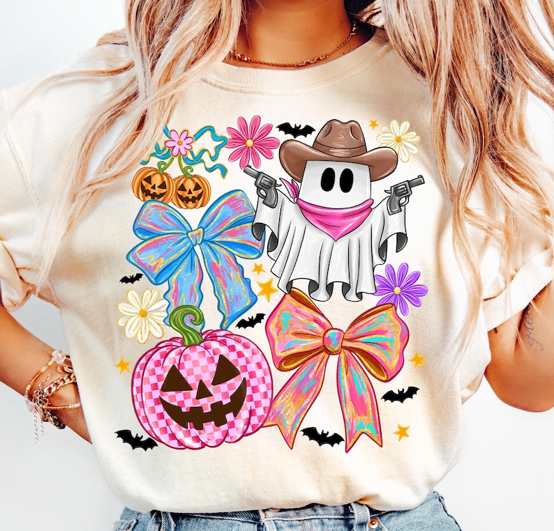 May include: A cream-coloured t-shirt with a Halloween design. The graphic features a ghost in a cowboy hat holding guns, alongside pumpkins, bows, flowers, bats, and stars.