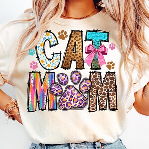 May include: A white t-shirt with the words "CAT MOM" in colorful, patterned letters. The letters are decorated with animal prints, paw prints, and a pink bow.