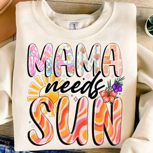Op de afbeelding: Een crèmekleurig sweatshirt met de tekst "Mama needs sun" in zwarte en kleurrijke letters. De letters zijn versierd met verschillende patronen, waaronder bloemen, golven en geometrische vormen. De zon is afgebeeld met een gele cirkel en oranje stralen.