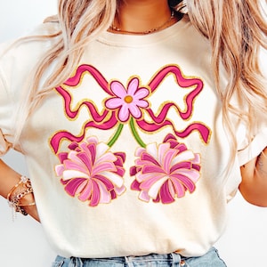 May include: Cream-colored sweatshirt featuring a pink bow and flower design with gold glitter accents. Below the bow are two pink and white pom-poms. The design is centered on the front of the shirt.