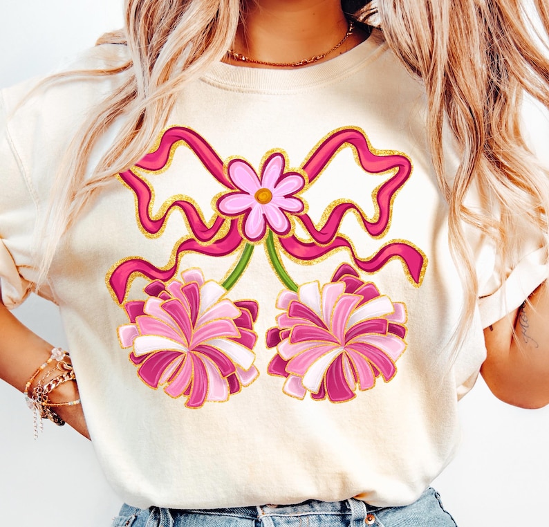 May include: Cream-colored sweatshirt featuring a pink bow and flower design with gold glitter accents. Below the bow are two pink and white pom-poms. The design is centered on the front of the shirt.