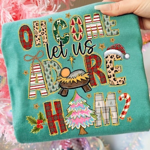 Pu&ograve; includere: T-shirt natalizia turchese con la frase "Oh Come Let Us Adore Him". Il design include un presepe, un albero di Natale e un bastoncino di zucchero. Le lettere presentano vari motivi, tra cui scozzese e animalier.