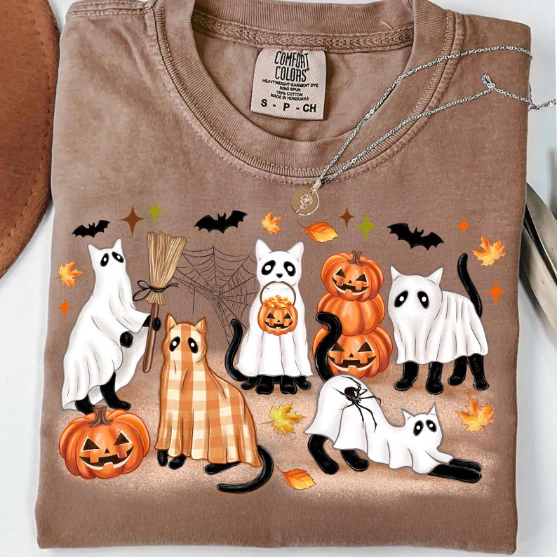 May include: Brown Comfort Colors t-shirt with a Halloween design. The graphic shows cartoon cats in ghost costumes, pumpkins, and bats. The shirt has a crew neck and short sleeves. A silver necklace is draped across the shirt.