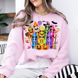 May include: A light pink sweatshirt featuring a colorful Halloween-themed crayon design. The design includes smiling crayons dressed as Halloween characters, pumpkins, ghosts, and bats. The word "Crayon" is visible on some crayons.