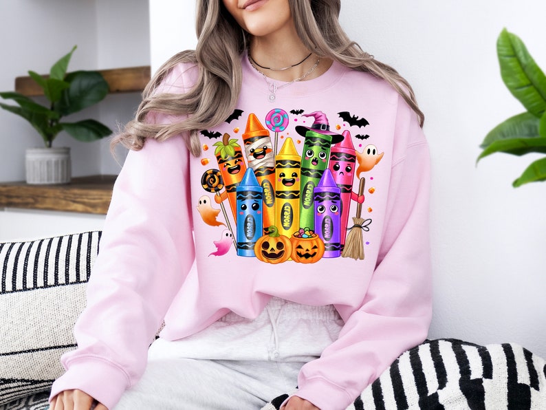 May include: A light pink sweatshirt featuring a colorful Halloween-themed crayon design. The design includes smiling crayons dressed as Halloween characters, pumpkins, ghosts, and bats. The word "Crayon" is visible on some crayons.