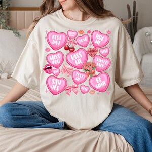 May include: A white t-shirt with a pink and white design of hearts with different phrases on them. The phrases include "Best Day", "Miss You", "Kiss Me", "Hug Me", "Sweet Talk", "Say Yes", "Be Mine", "Text Me", and "XOXO".