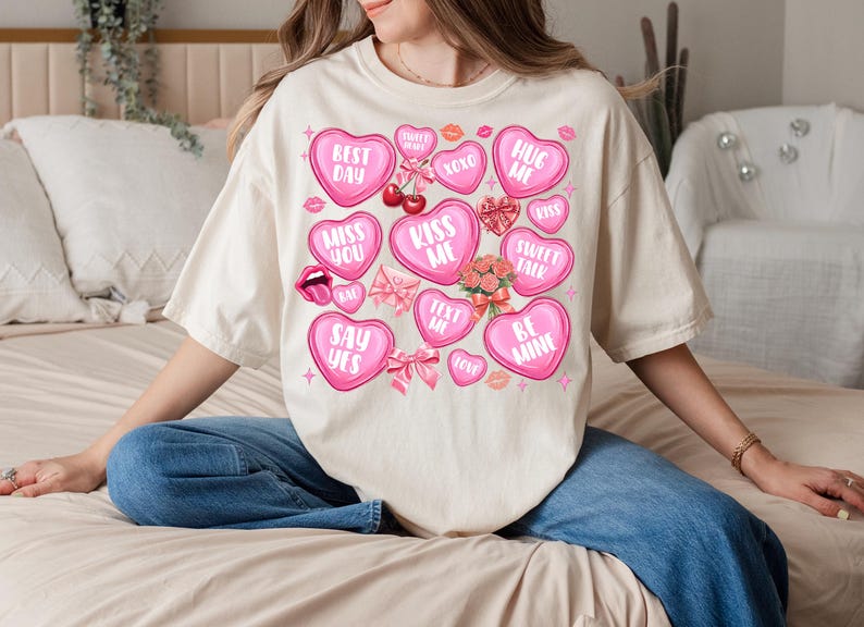 May include: A white t-shirt with a pink and white design of hearts with different phrases on them. The phrases include "Best Day", "Miss You", "Kiss Me", "Hug Me", "Sweet Talk", "Say Yes", "Be Mine", "Text Me", and "XOXO".