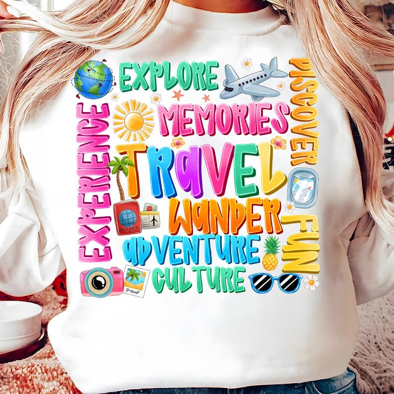 May include: White sweatshirt with a colorful graphic design featuring the words "Explore", "Memories", "Travel", "Wander", "Adventure", "Culture", "Discover", "Fun", and "Experience". The graphic also includes images of a globe, airplane, palm trees, camera, sunglasses, pineapple, and flowers.