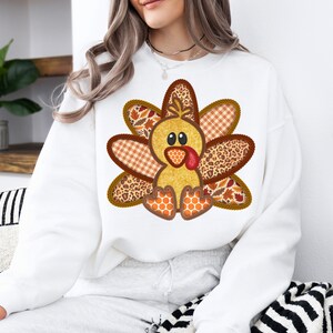 May include: White sweatshirt featuring a charming patchwork turkey design. The turkey is brown, yellow and orange with a red wattle. The design is ideal for Thanksgiving.