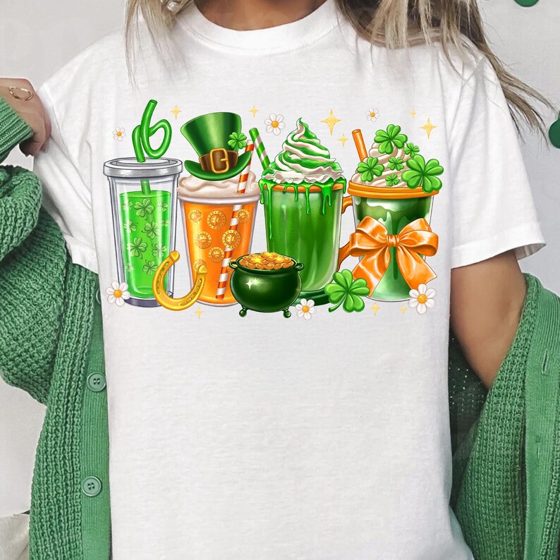 May include: White t-shirt with a graphic of four green, orange, and white drinks with shamrocks and a gold pot of gold. The drinks are topped with whipped cream, a green shamrock, and a leprechaun hat.