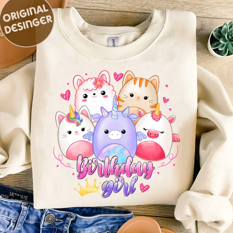 Puede incluir: Una sudadera de color crema con un gr&aacute;fico de cinco animales de dibujos animados, incluyendo un unicornio, un gato, una llama y dos criaturas parecidas a cerdos. Los animales est&aacute;n rodeados de corazones rosas y el texto "Birthday girl" con una corona dorada.