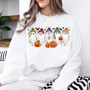 Op de afbeelding: Wit sweatshirt met een afbeelding van vijf geesten die kleurrijke hoeden dragen en pompoenen en koffiebekers vasthouden. De geesten staan in een rij en hebben een schattige cartoon-stijl.