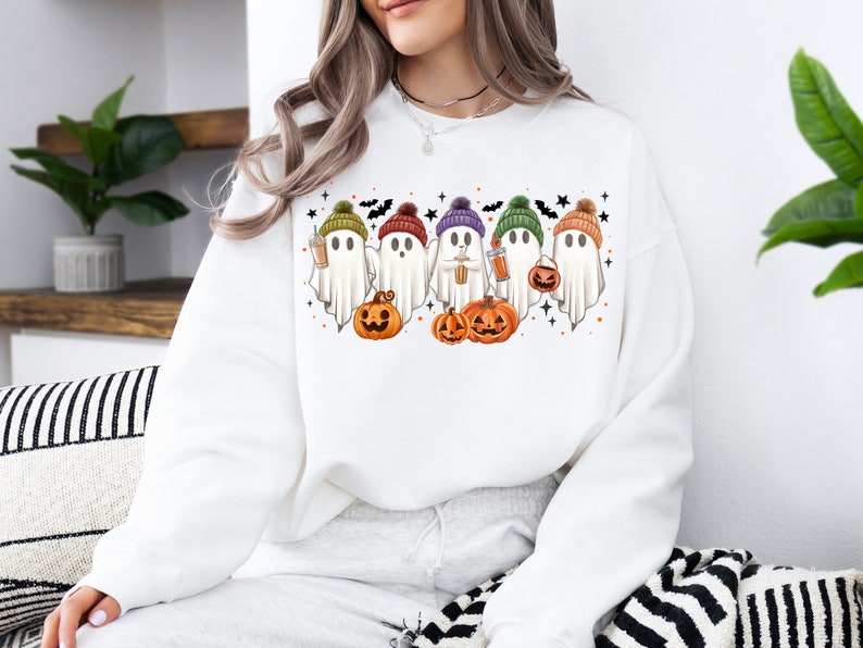 Op de afbeelding: Wit sweatshirt met een afbeelding van vijf geesten die kleurrijke hoeden dragen en pompoenen en koffiebekers vasthouden. De geesten staan in een rij en hebben een schattige cartoon-stijl.
