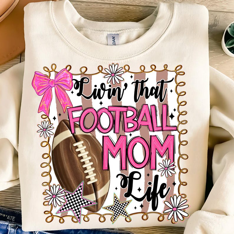 May include: Cream-colored sweatshirt with a graphic that reads "Livin' that FOOTBALL MOM Life" in pink and black. The design includes a football, a pink bow, daisies, and checkered stars. The sweatshirt is a casual style.