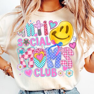 May include: A white t-shirt with a colorful graphic design that says "Anti Social Mom's Club". The design features a smiley face, stars, hearts, and checkered patterns.